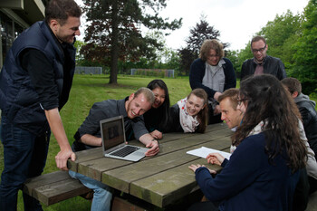 Größere Gruppe von Studierenden arbeitet gemeinsam im Freien und ist über einen Laptop gebeugt