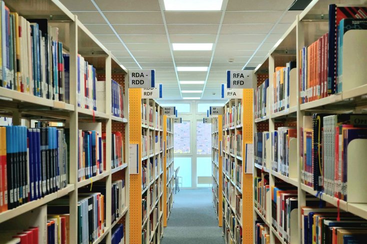 Abgebildet wird der Lesesaal der Hochschulbibliothek Wilhelmshaven mit einem schmalen Gang zwischen hohen, hellen Metallregalen. In den Regalen stehen zahlreiche Bücher. 