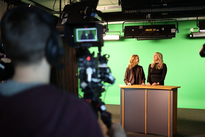 Raumansicht des Videostudios mit Moderationstisch, an dem zwei Studentinnen stehen, und Greenscreen. Unscharf im Vordergrund ist der Kameramann zu sehen.