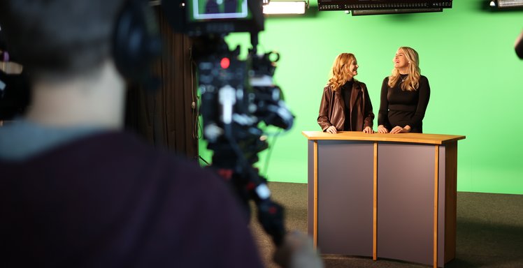 Raumansicht des Videostudios mit Moderationstisch, an dem zwei Studentinnen stehen, und Greenscreen. Unscharf im Vordergrund ist der Kameramann zu sehen.