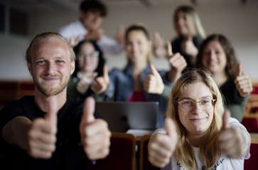 Sieben Studierende recken in einem Hörsaal lachend beide Daumen hoch.