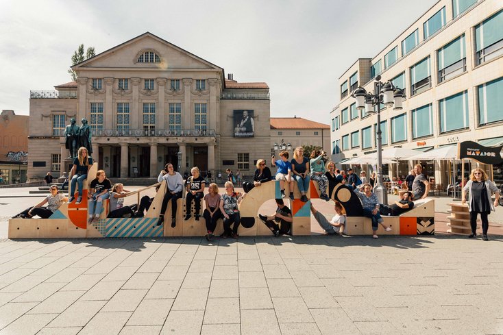 Begehbare Holzinstallation mit geometrischen Formen auf einem städtischen Platz; zahlreiche Menschen sitzen davor, dahinter ein historisches Gebäude.