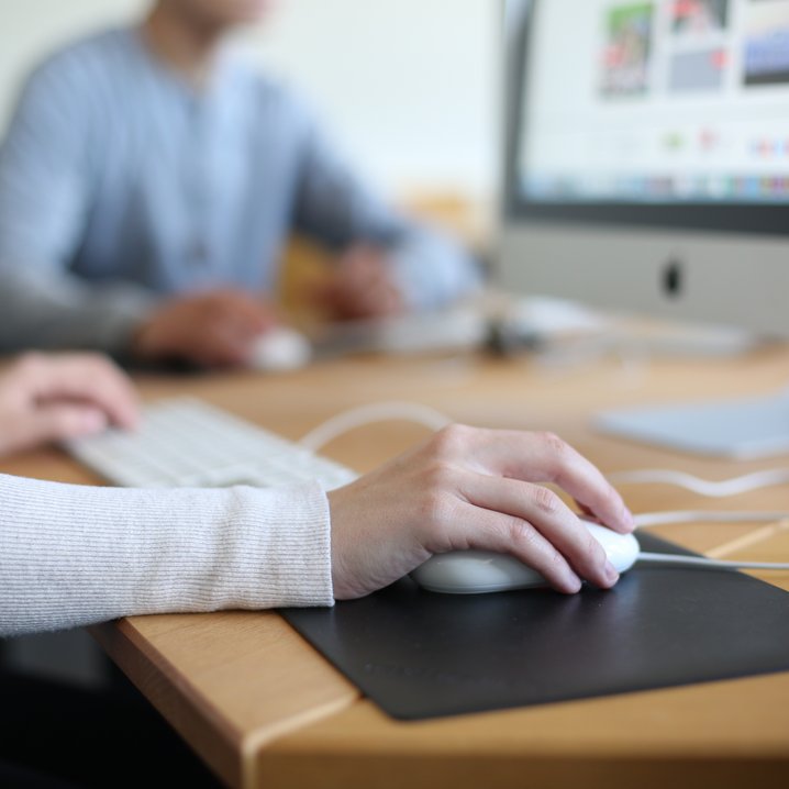 Die Nahaufnahme fokussiert auf eine Hand, die eine Computermaus auf einem dunklen Pad bedient, während im Hintergrund eine weitere Person unscharf an einem Monitor arbeitet. Die Szene verdeutlicht die Nutzung moderner IT-Arbeitsplätze in einer hellen Lernumgebung.