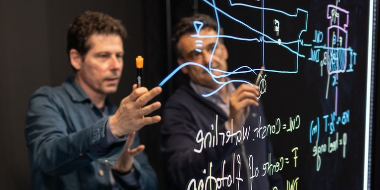 Zwei Professoren stehen hinter dem Lightboard und diskutieren über die Zeichnung, die auf dem Lightboard zu sehen ist.