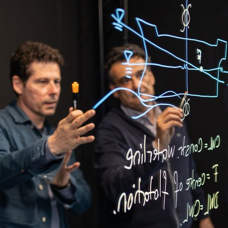 Zwei Professoren stehen hinter dem Lightboard und diskutieren über die Zeichnung, die auf dem Lightboard zu sehen ist.