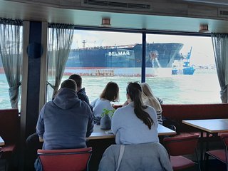 Studierende machen eine Hafenrundfahrt. Im Hintergrund fährt ein Containerschiff vorbei.