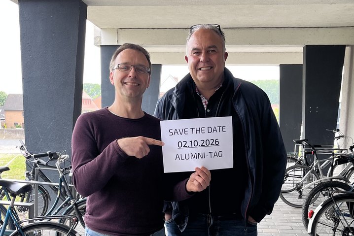 Zwei freundlich lächelnde Mitarbeitende des Fachbereichs Seefahrt und Logistik stehen nebeneinander und halten ein Schild hoch, auf dem der Text „Save the date 02.10.2026 Alumni-Tag“ zu sehen ist.