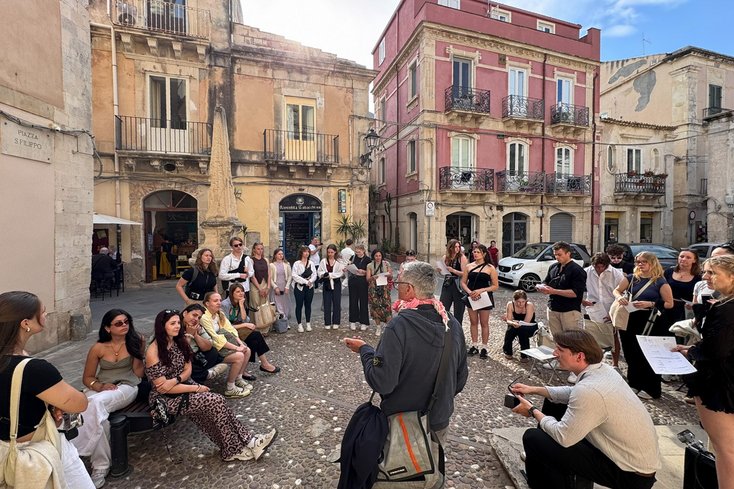 Studierende während einer Exkursion in Italien sitzen auf einem Marktplatz und hörem einem Dozenten zu