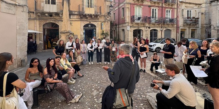 Studierende während einer Exkursion in Italien sitzen auf einem Marktplatz und hörem einem Dozenten zu