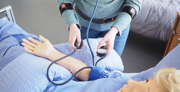 Eine Person ist mit Sensoren ausgestattet und versorgt eine Patientin im Krankenhausbett, die durch eine Puppe dargestellt wird.