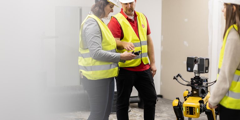 Zwei Studierende und ein Dozent befinden sich mit einem Roboterhund auf der Baustelle im Inneren eines Gebäudes.