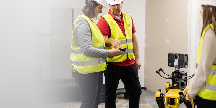 Zwei Studierende und ein Dozent befinden sich mit einem Roboterhund auf der Baustelle im Inneren eines Gebäudes.