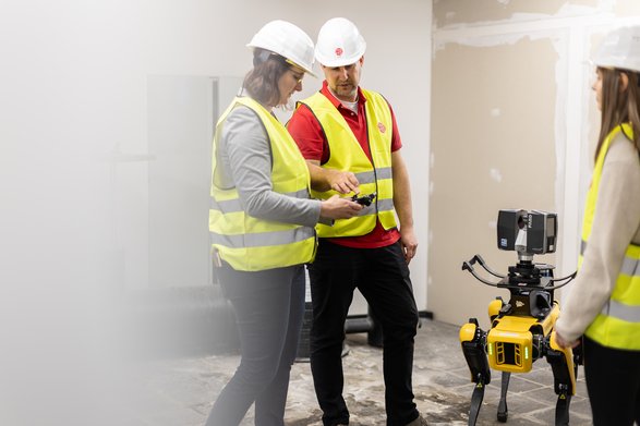 Zwei Studierende und ein Dozent befinden sich mit einem Roboterhund auf der Baustelle im Inneren eines Gebäudes.