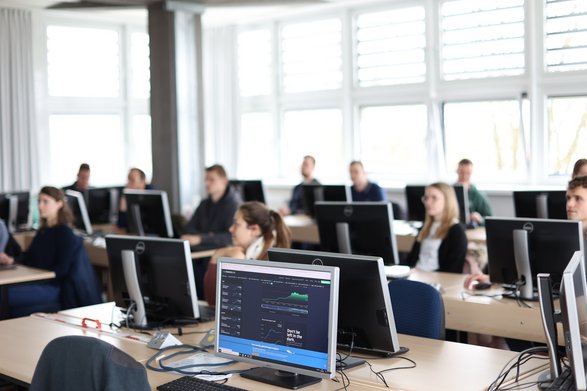 In einem Vorlesungsraum sitzen 12 Studierende an Rechnern und hören dem Dozenten zu.