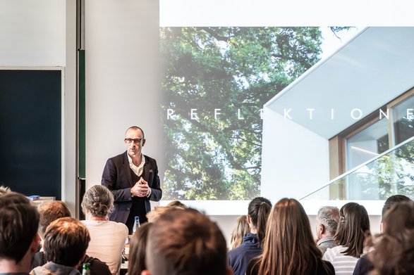 Ein Mann hät einen Vortrag in einem Hörsaal im Hintergrund eine Präsentation mit Architektur