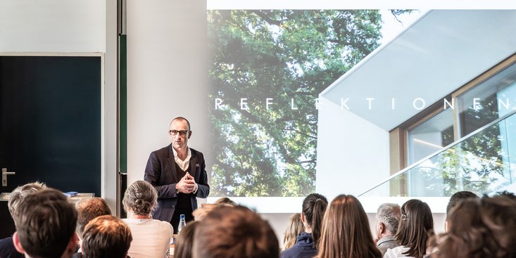 Ein Mann hät einen Vortrag in einem Hörsaal im Hintergrund eine Präsentation mit Architektur