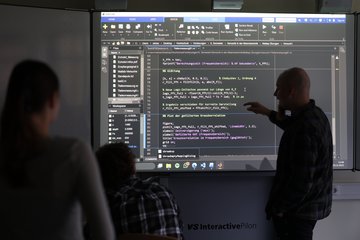 In einem dunklen Raum werden Programmiercodes an einem Smartboard erklärt.