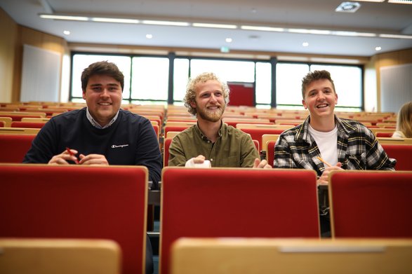 Drei Studierende sitzen im Vorlesungssaal und lächeln in die Kamera.