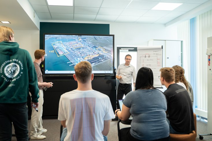 Ein Dozent erklärt auf einem Bildschirm ein Hafengelände. Studierende hören zu.