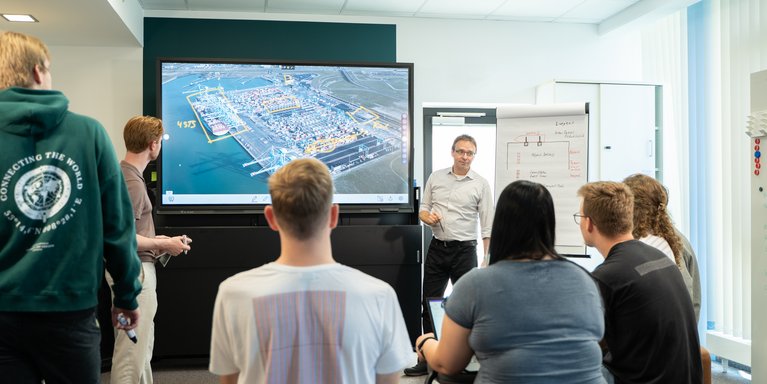 Ein Dozent erklärt auf einem Bildschirm ein Hafengelände. Studierende hören zu.