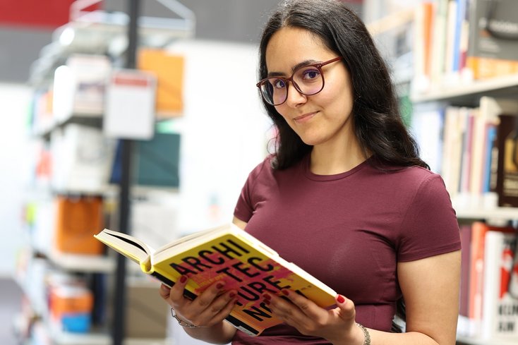 Eine Studentin steht mit einem Buch neben einem Bücherregal und schaut in die Kamera