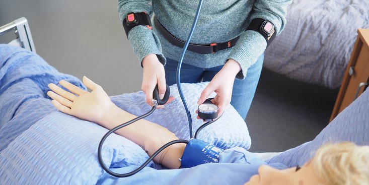 Eine Person ist mit Sensoren ausgestattet und versorgt eine Patientin im Krankenhausbett, die durch eine Puppe dargestellt wird.