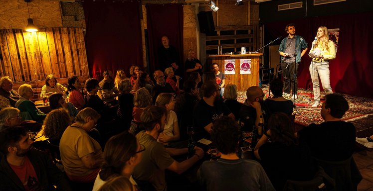 Zwei Personen stehen in einer Kneipe auf der Bühne. Vor ihnen sitzen viele Zuhörerinnen und Zuhörer.