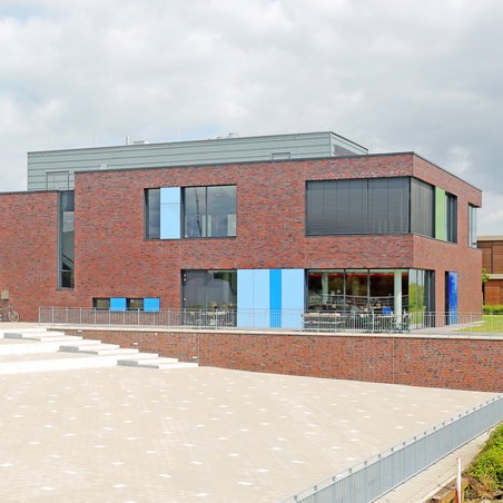 Das moderne Bibliotheksgebäude am Campus Elsfleth besticht durch eine rote Backsteinfassade, die durch bunte Paneele in Blau- und Grüntönen an den Fenstern aufgelockert wird. Im Vordergrund erstreckt sich ein großer, gepflasterter Platz mit flachen Stufen, der direkt zum Eingang des zweistöckigen Gebäudes führt.