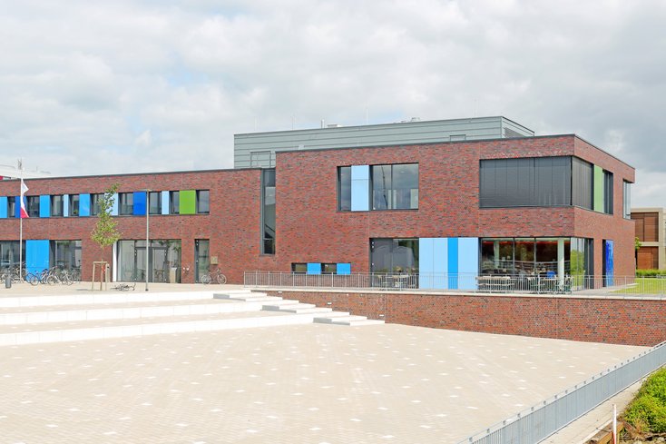 Das moderne Bibliotheksgebäude am Campus Elsfleth besticht durch eine rote Backsteinfassade, die durch bunte Paneele in Blau- und Grüntönen an den Fenstern aufgelockert wird. Im Vordergrund erstreckt sich ein großer, gepflasterter Platz mit flachen Stufen, der direkt zum Eingang des zweistöckigen Gebäudes führt.
