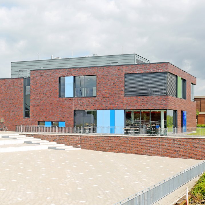 The modern library building at Elsfleth Campus has a red brick exterior, speckled with blue and green panels next to the windows. A big paved square with a wide staircase leads to the entrance of the two-story building.