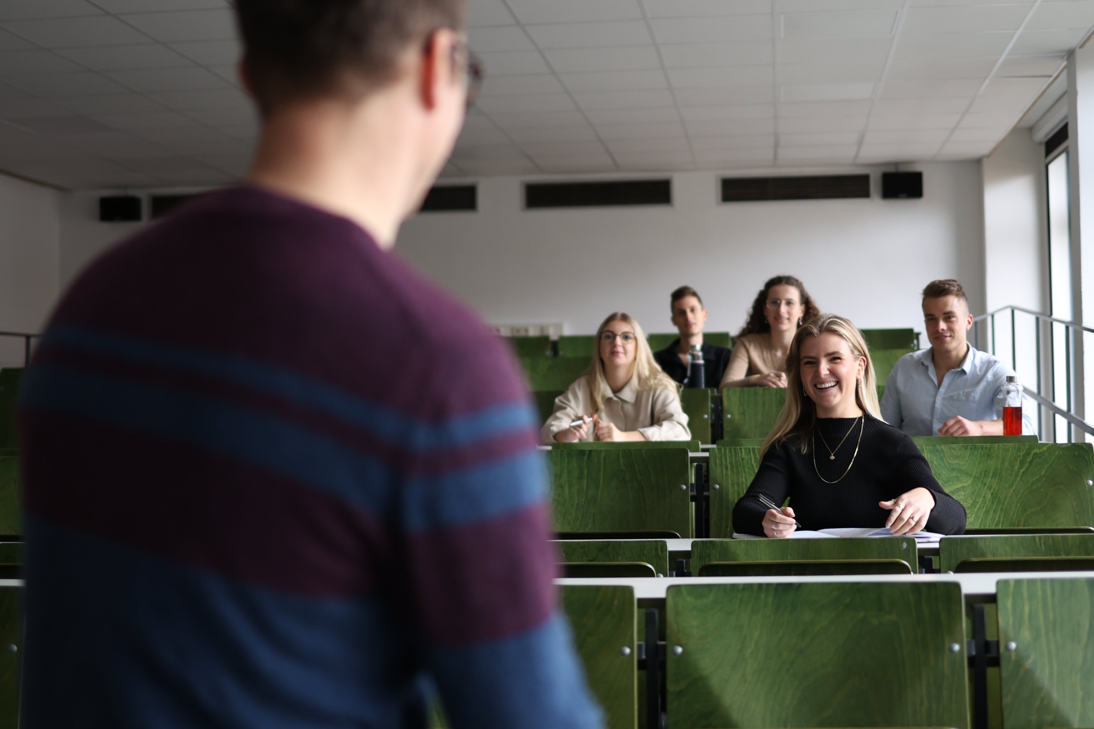 Dozent sitzt locker auf dem Dozententisch und erklärt Studierenden im Vorlesungssaal etwas.