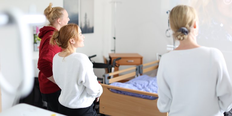 Drei Studierende stehen vor einem Pflegebett und schauen auf eine Präsentation an der Wand.