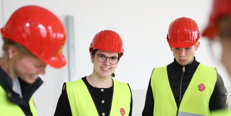 Studierende stehen auf einer Baustelle drinnen mit Helmen und Weste.