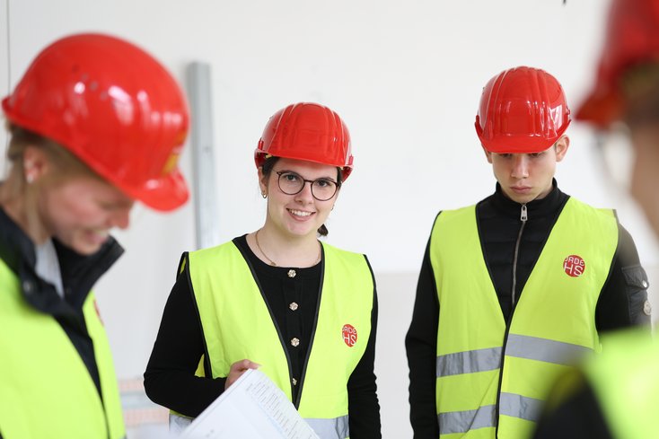 Studierende stehen auf einer Baustelle drinnen mit Helmen und Weste.