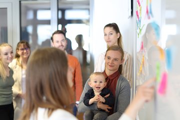Gruppe Studierender löst Aufgabe am Whiteboard mit Post-Its, eine Studentin hat ein Baby auf dem Arm.