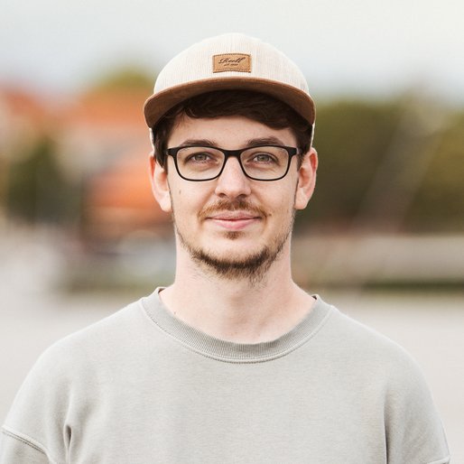 Portraitbild eines jungen Mannes mit Brille und Basecap, der in die Kamera lächelt