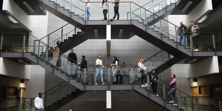 Mehrere junge Menschen stehen verteilt auf einer symmetrischen, modernen Treppenanlage in einem hellen, mehrstöckigen Atrium. Die Personen unterhalten sich in kleinen Gruppen auf verschiedenen Ebenen und Treppenabsätzen, während die Architektur durch klare Linien und Glasgeländer geprägt ist.