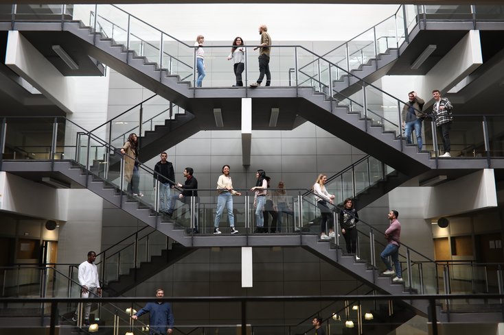 Mehrere junge Menschen stehen verteilt auf einer symmetrischen, modernen Treppenanlage in einem hellen, mehrstöckigen Atrium. Die Personen unterhalten sich in kleinen Gruppen auf verschiedenen Ebenen und Treppenabsätzen, während die Architektur durch klare Linien und Glasgeländer geprägt ist.