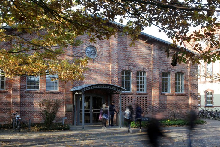 Die Aufnahme zeigt den Eingangsbereich einer Bibliothek in einem historischen Backsteingebäude mit Rundbogenfenstern und einem kleinen Vordach über der Tür. Im Vordergrund sind schemenhaft Passanten in Bewegung zu sehen, während herbstliche Bäume das Gebäude einrahmen.