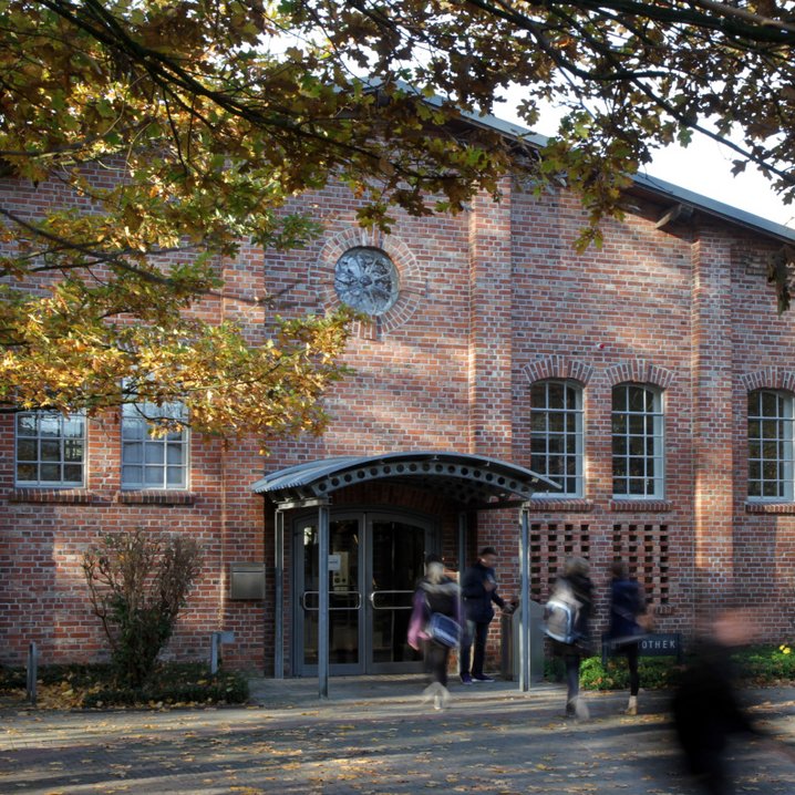 The entrance of Oldenburg Library, a historic brick building with round arch windows and a small roof above the door. People walking in and out are pictured out of focus, and the building is framed by autumnal trees.