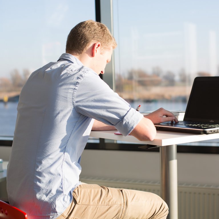 Ein junger Mann sitzt an einem hohen Tisch vor einem Laptop und macht sich Notizen in ein Heft. Durch die große Fensterfront im Hintergrund fällt helles Sonnenlicht auf seinen Arbeitsplatz und bietet einen Ausblick auf eine Wasserfläche und Bäume.