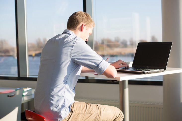 Ein junger Mann sitzt an einem hohen Tisch vor einem Laptop und macht sich Notizen in ein Heft. Durch die große Fensterfront im Hintergrund fällt helles Sonnenlicht auf seinen Arbeitsplatz und bietet einen Ausblick auf eine Wasserfläche und Bäume.