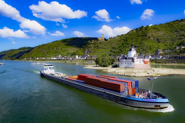 Luftaufnahme des Rheins mit einem Binnenschiff, einem Dorf am Ufer und einer Burg. 