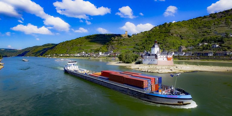 Luftaufnahme des Rheins mit einem Binnenschiff, einem Dorf am Ufer und einer Burg. 