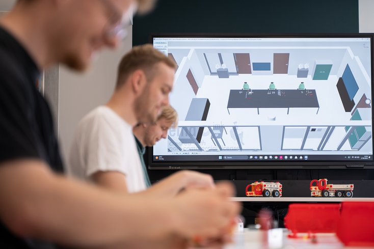 Drei junge Menschen arbeiten im Logistiklabor vor einem großen Bildschirm