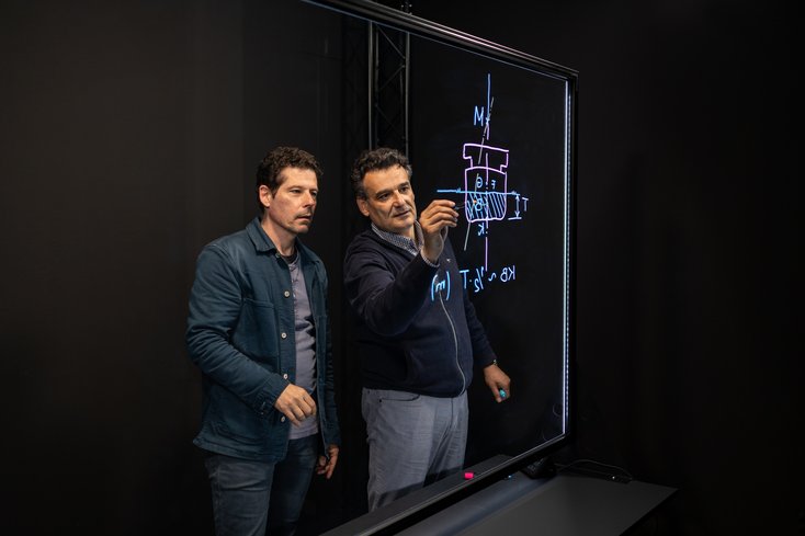 Zwei Dozenten stehen vor dem Lightboard. Einer davon zeichnet etwas auf das Lightboard.