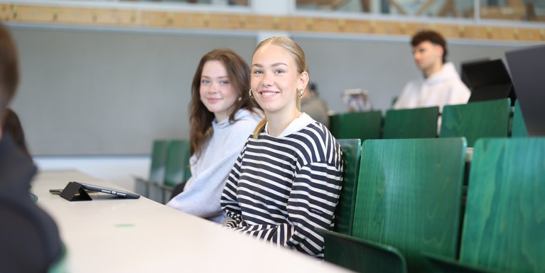 Zwei Studentinnen sitzen im Vorlesungssaal und lächeln in die Kamera.