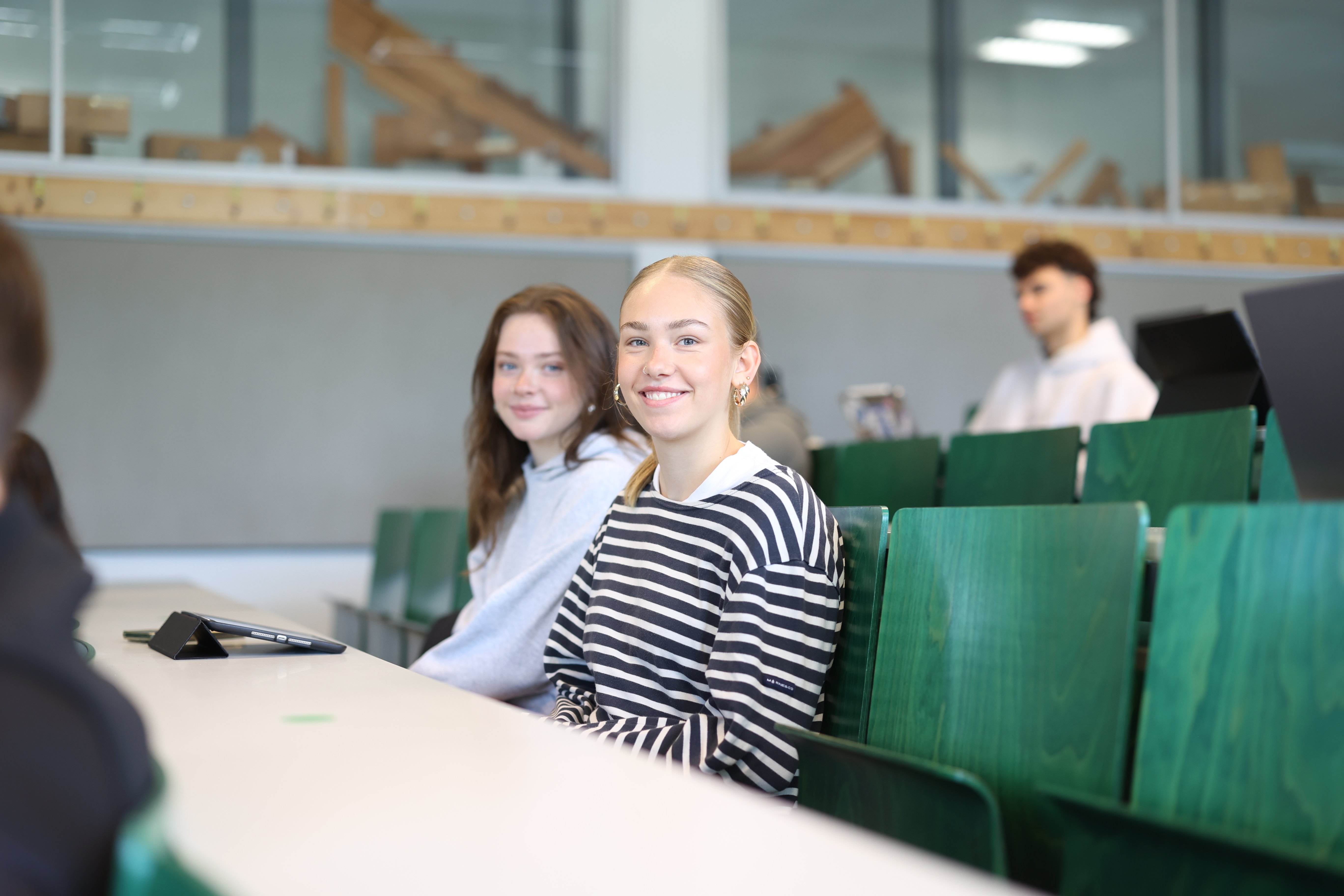 Zwei Studentinnen sitzen im Vorlesungssaal und lächeln in die Kamera.