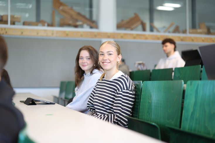 Zwei Studentinnen sitzen im Vorlesungssaal und lächeln in die Kamera.