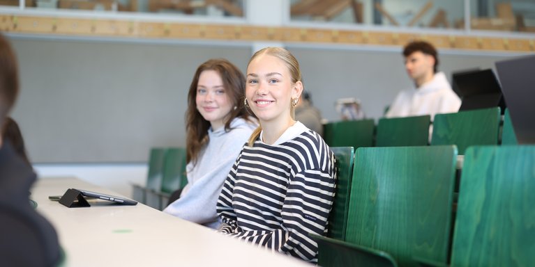 Zwei Studentinnen sitzen im Vorlesungssaal und lächeln in die Kamera.